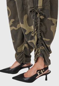 Gambe in pantaloni arricciati camouflage abbinati a tacchi a punta neri con cinturino alla caviglia e dettagli in oro.