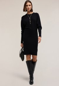 Abito lungo nero a maniche lunghe con silhouette aderente, abbinato a stivali neri attillati fino al ginocchio e una piccola borsa nera. Design semplice ed elegante.