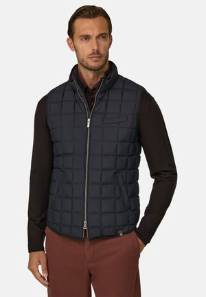 Homme portant une veste sans manches noire matelassée par-dessus une chemise sombre à manches longues et un pantalon marron, regardant sur le côté avec les mains le long du corps.