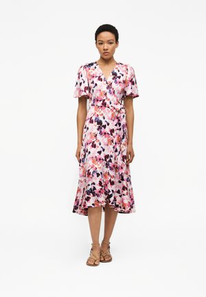 Γυναίκα όρθια, φοράει ένα ροζ φόρεμα τύπου wrap με λουλουδένιο μοτίβο, με κοντά μανίκια που φουσκώνουν και μπεζ σανδάλια με λουράκια, σε απλό λευκό φόντο.