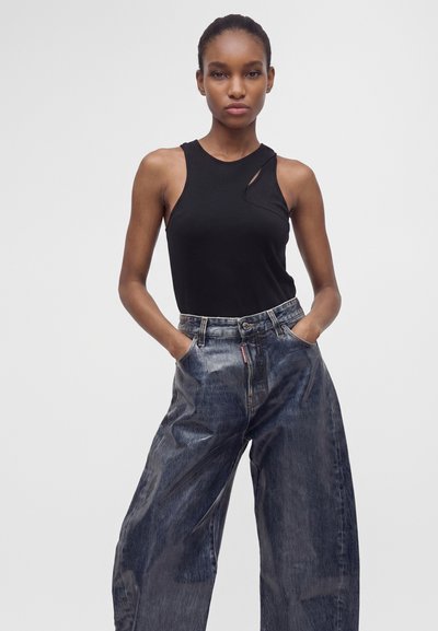 Jeune femme portant un haut noir sans manches avec un détail découpé et un jean bleu délavé et ample, debout les mains dans les poches devant un fond uni.
