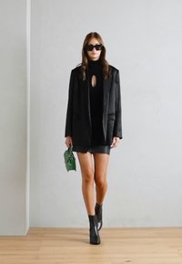Blazer preto sobre uma blusa preta com recorte em forma de olho-chave, saia mini de couro preta e botas de tornozelo pretas, segurando uma pequena bolsa verde.