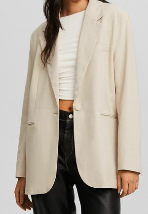 Kobieta ubrana w beżową marynarkę na jeden guzik, nałożoną na biały krótki top oraz czarne skórzane spodnie z wysokim stanem, stoi z rozluźnionymi ramionami.