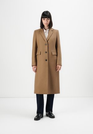 Vrouw staand in een lange camelkleurige jas met zwarte knopen, donkere broek, zwarte instappers en een lichtgekleurde overhemd met kraag tegen een witte achtergrond.