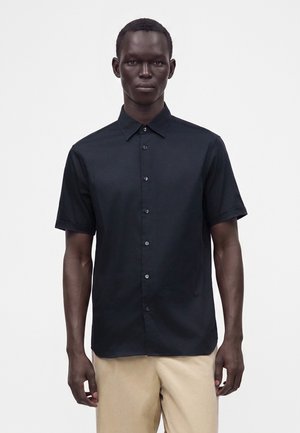 Homme portant une chemise noire à manches courtes boutonnée et un pantalon beige, debout devant un fond clair uni.