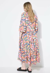 Frau mit langen blonden Haaren, die ein farbenfrohes Midikleid mit Bananenmuster und schwarze Clogs trägt, steht vor einem weißen Hintergrund.