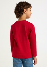 Langærmet rød bomuldsbluse med rund hals, der har en glat tekstur og et enkelt design, båret med blå denimbukser.