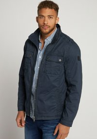 Marineblauwe ritsjack met een staande kraag, twee voorzakken, gestructureerde stof en stiksels. Draag het over een gestreept shirt.