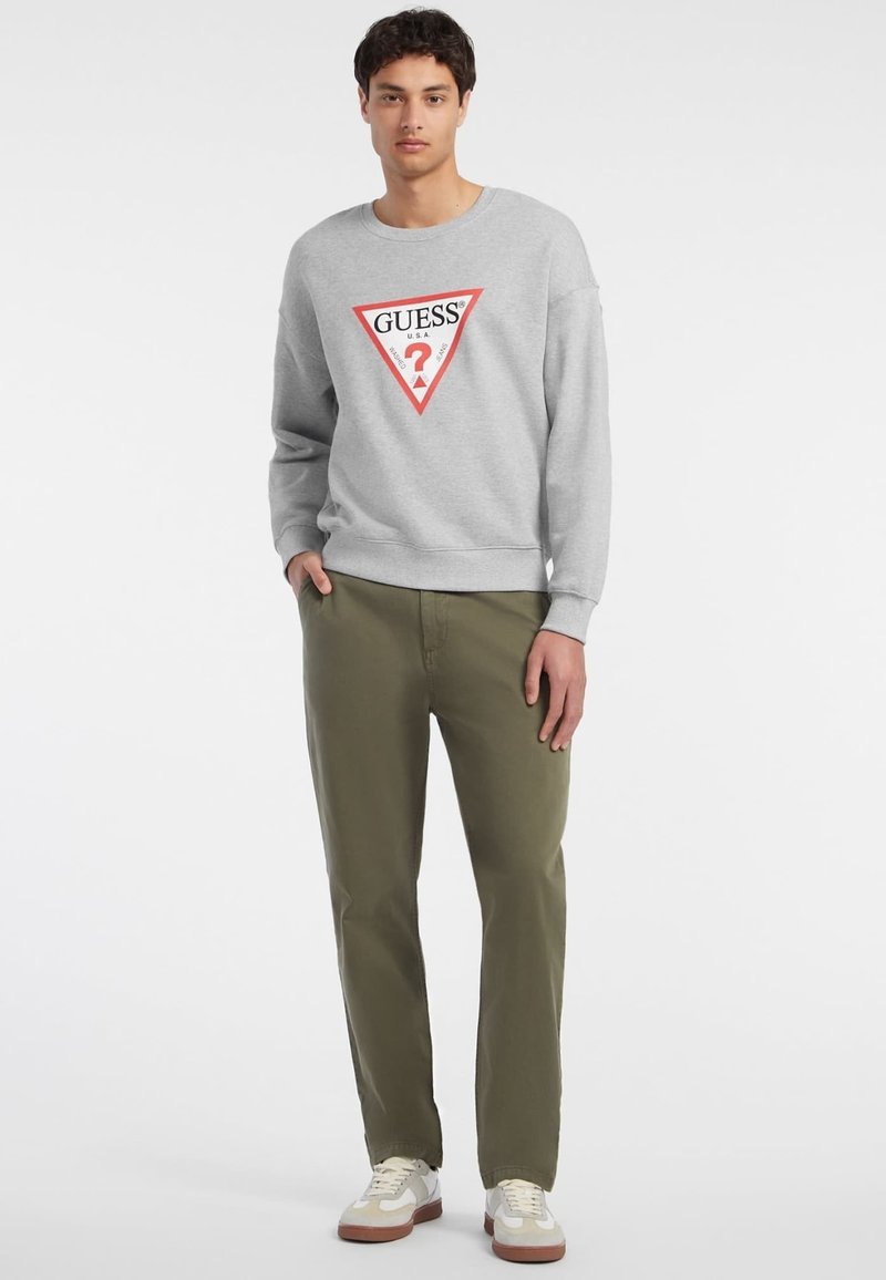 Felpa grigia con un logo triangolare rosso e bianco, abbinata a pantaloni verde oliva. L'outfit presenta un design casual con polsini a coste.