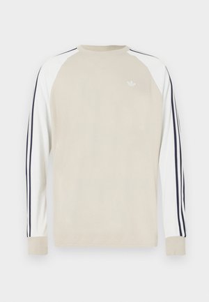 Béžové tričko s dlouhým rukávem, bílé raglánové rukávy a námořnicky modré pruhy podél stran. Na hrudi je malé bílé logo.