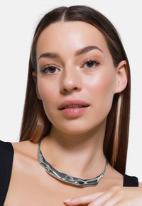 Collana di metallo elegante con un design ondulato, rifinita in argento lucido. Texture liscia con riflessi sottili; forma minimalista e contemporanea.