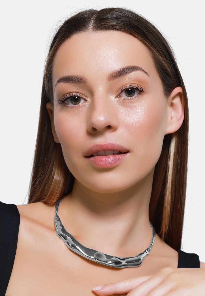 Collana di metallo elegante con un design ondulato, rifinita in argento lucido. Texture liscia con riflessi sottili; forma minimalista e contemporanea.