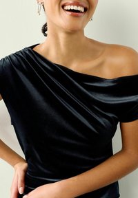 Sort off-the-shoulder top i blødt, skinnende stof med draperet detalje. Har korte ærmer og en tætsiddende design, der afslører skulder og kraveben.