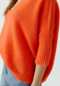 Übergrößter orangefarbener Strickpullover mit V-Ausschnitt, strukturierten kurzen Ärmeln und einer weichen, flauschigen Textur. Der Saum ist am unteren Ende geschwungen.