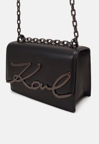 Borsa a tracolla strutturata nera con esterno in pelle liscia. Presenta una tracolla a catena e un grande logo metallico "Karl" sul fronte.