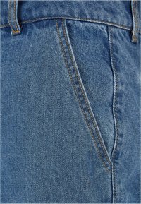 Jeans de mezclilla de color azul claro, con costuras visibles en naranja contrastante. Primer plano del área del bolsillo frontal y la textura.