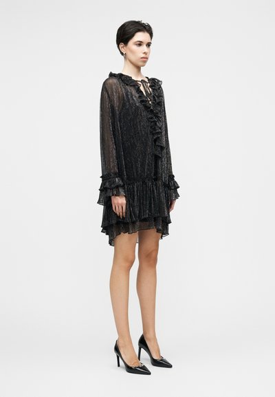 Μαύρο φόρεμα με μακριά μανίκια, βολάν, διάφανο ύφασμα και στρώσεις στο τελείωμα. Συνδυάζεται με μυτερές μαύρες γόβες, αναδεικνύοντας μια κομψή σιλουέτα.