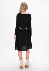Rochie neagră cu mâneci lungi și talie strânsă, având o margine de culoare deschisă în jurul decolteului. Purtată cu cizme cowboy bej.