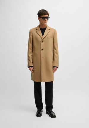 Manteau en mélange de laine beige avec un col cranté, trois boutons, une poche poitrine et des poches latérales, associé à un pantalon noir et des chaussures noires.