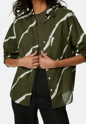 Camicia verde con bottoni, motivo a onde bianche, tessuto strutturato e taschino sul petto. Abbinata a una parte superiore scura e pantaloni neri.