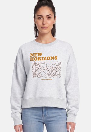 Femme portant un sweat-shirt gris clair avec "New Horizons" et un dessin au trait d'une scène de randonnée en marron, associé à un jean bleu.