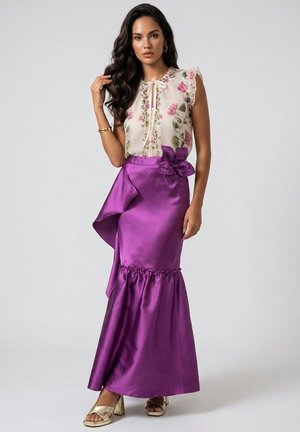 Mujer que lleva una blusa sin mangas con estampado floral y una falda larga y brillante de color púrpura con un gran detalle de flor en la cintura, posando sobre un fondo liso.