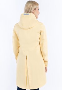 Impermeabil galben cu glugă, proiectat cu un pli ant în spate și o textură netedă. Are un accent cu dungi pe interiorul glugii.