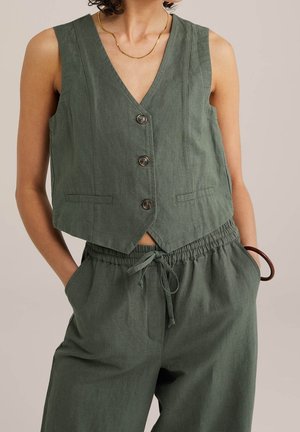 Mouwloze olijfgroene gilet met knopen, gecombineerd met bijpassende broek met koord, gedragen door een persoon met handen in de zakken en een gouden ketting.