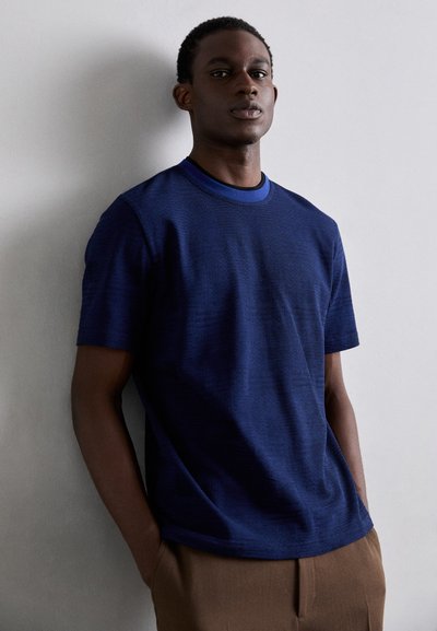 Mørkeblå T-skjorte med kortermet i teksturert stoff, med rund hals og subtilt mønster; kombinert med lysebrune bukser.
