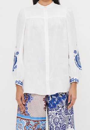 Femme portant un chemisier blanc boutonné avec une broderie florale bleue sur les manches et un pantalon à motifs multicolores avec des dessins bleus et marron.