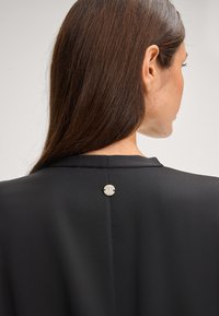 Czarna marynarka o gładkiej fakturze, z akcentem w srebrnym kolorze na plecach w postaci okrągłego wzoru. Prosta i elegancka sylwetka.