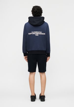 Personne aux cheveux bouclés, vue de dos, portant un sweat à capuche bleu marine avec le texte "EMPORIO ARMANI 1981" et un short noir avec des chaussures noires.
