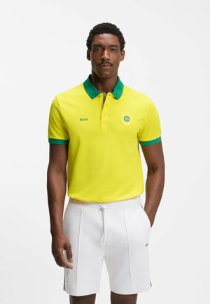 Man draagt een geel poloshirt met groene kraag en mouwen, witte korte broek, staand tegen een effen witte achtergrond.