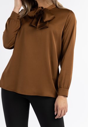 Femme portant un chemisier en satin marron à manches longues avec un col lavallière et un pantalon noir cintré, debout devant un fond uni.