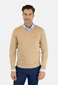 Suéter de punto suave en color beige claro con escote en forma de V, con puños y dobladillo de canalé, combinado con una camisa de cuello azul claro debajo.