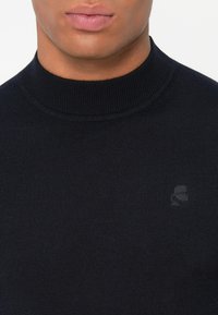 Granatowy sweter z wysokim prążkowanym kołnierzem, wykonany z miękkiego materiału. Posiada małe, tonalne logo na lewej piersi. Gładka tekstura.
