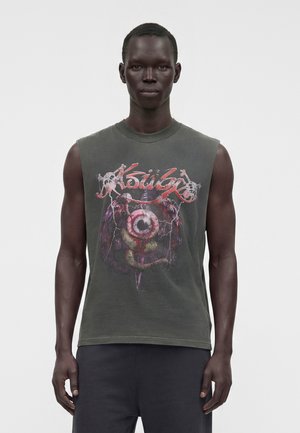 Jeune homme à la peau foncée portant un t-shirt graphique sans manches gris foncé avec un motif abstrait rouge et blanc et un pantalon noir, debout devant un fond uni.