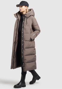 Long brown puffer coat with a hood, zippered front, and large pockets; worn over a black outfit and paired with black ankle boots.