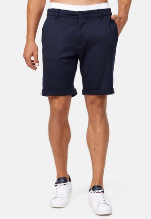 Pantalones cortos de algodón azul marino con frente plano, dobladillos enrollados y bolsillos laterales. Cuenta con un cierre de botón y trabillas para cinturón. Llevado con zapatillas blancas.
