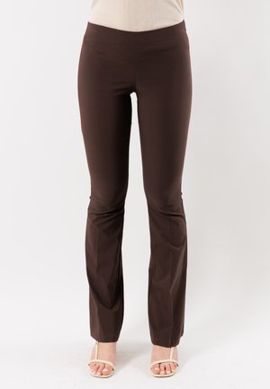 Pantaloni marroni aderenti indossati da una persona in piedi, abbinati a sandali aperti beige chiaro su uno sfondo semplice.