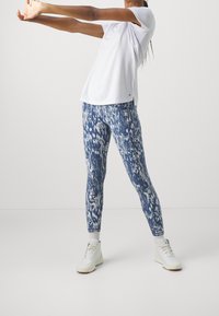 Vit kortärmad topp i kombination med blå mönstrade leggings med ormskinnsmönster. Modellen bär vita sneakers under en stretchpose.