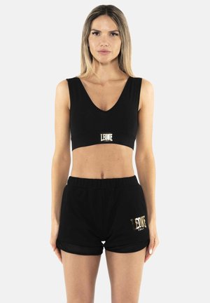 Donna in piedi, rivolta in avanti, indossa un top corto nero senza maniche e pantaloncini neri con loghi dorati "Leone 1947" su entrambi gli indumenti.