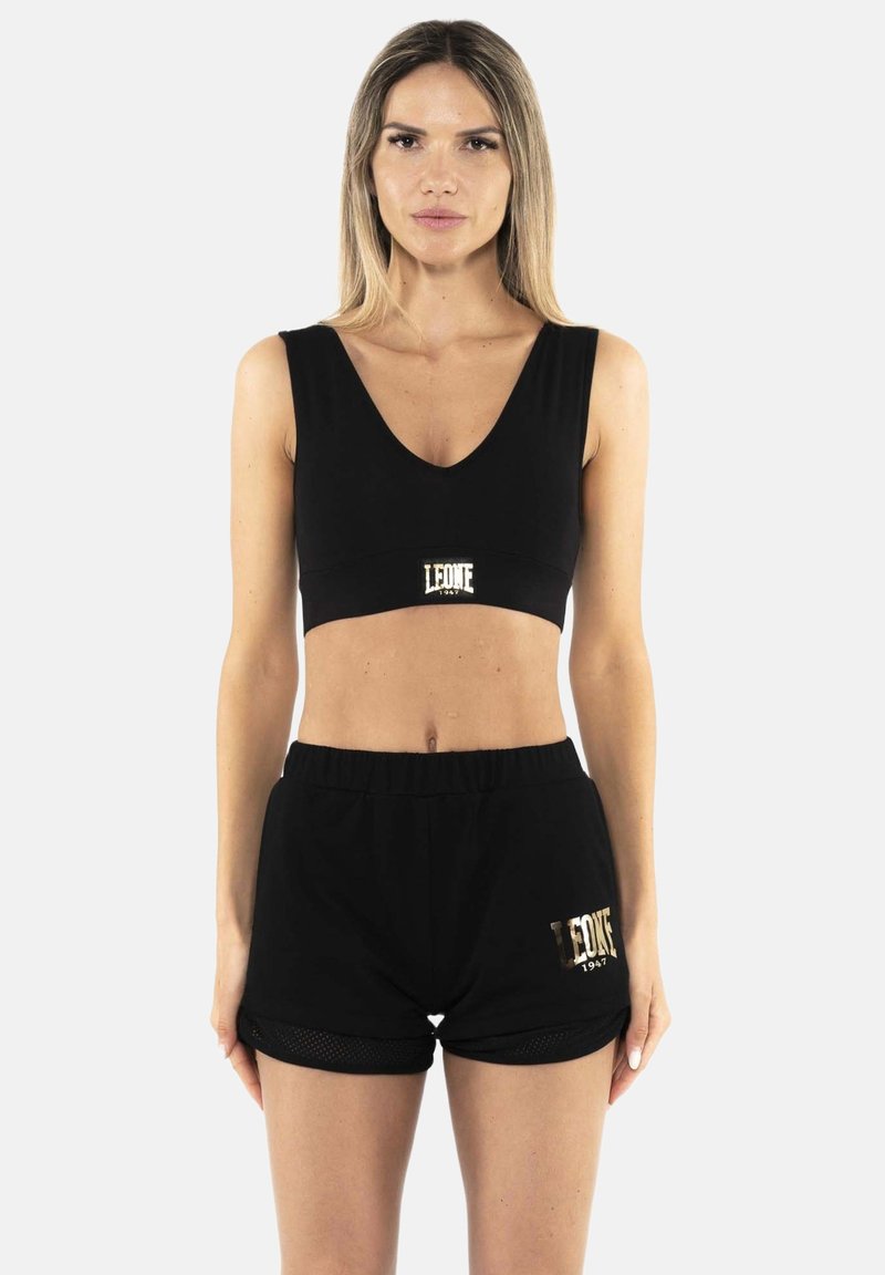 Femme debout face à l'avant portant un crop top noir sans manches et un short noir avec des logos dorés "Leone 1947" sur les deux vêtements.