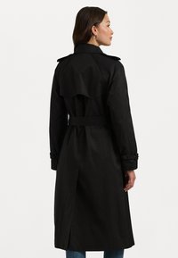 Musta trench-coat, jossa on kaksirivinen malli, vyöllä varustettu vyötärö ja keskipitkä leikkaus. Sisältää takaselän läpän ja hihnat, joissa on säädettävät remmit.