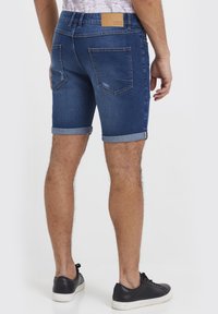 Denimshorts i mørk blå med oppbrettede kanter og subtile slitasjedetaljer. Har to baklommer og en lærlapp på linningen.