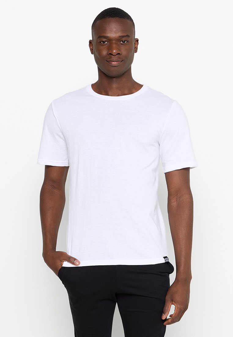 T-shirt en coton blanc, manches courtes, encolure ronde, coupe décontractée. Présente un branding minimal sur l'ourlet ; associé à un pantalon sombre.