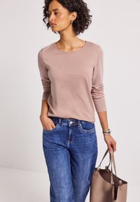 Femme avec des cheveux bouclés courts, portant une chemise mauve à manches longues et un jean bleu, tenant un sac fourre-tout beige et regardant sur le côté.