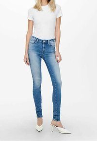 Vaqueros de mezclilla ajustados de color azul con un acabado desgastado, diseño de cinco bolsillos y cintura de altura media, combinados con una camiseta blanca sencilla y mulas blancas de tacón.