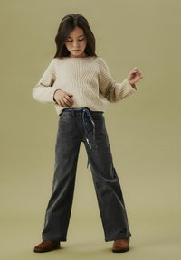 Beżowy sweter z prążkowanej dzianiny z długimi rękawami, zestawiony z szerokimi spodniami w ciemnoszarym kolorze, z niebieskim wzorowanym sznurkiem, oraz brązowymi botkami za kostkę.
