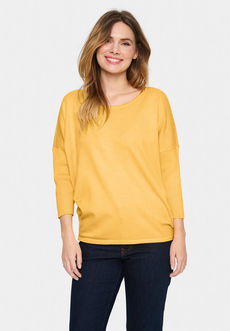Gul, löst sittande tröja med trekvartsärmar, ribbat mönster på ärmarna och rundad halsringning, i kombination med mörka jeans.